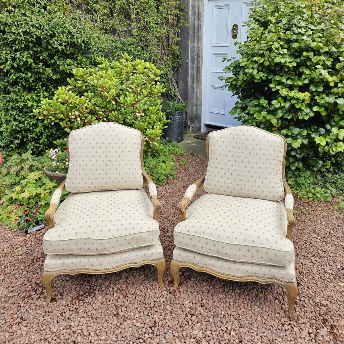 A pair of antique French-style bergère armchairs reupholstered in a cream fleur-de-lis fabric, set on gravel by a panelled door