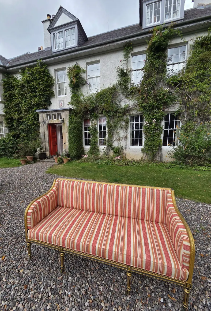 A gilt-frame settee reupholstered in a red and cream striped fabric, on the gravel outside a country house
