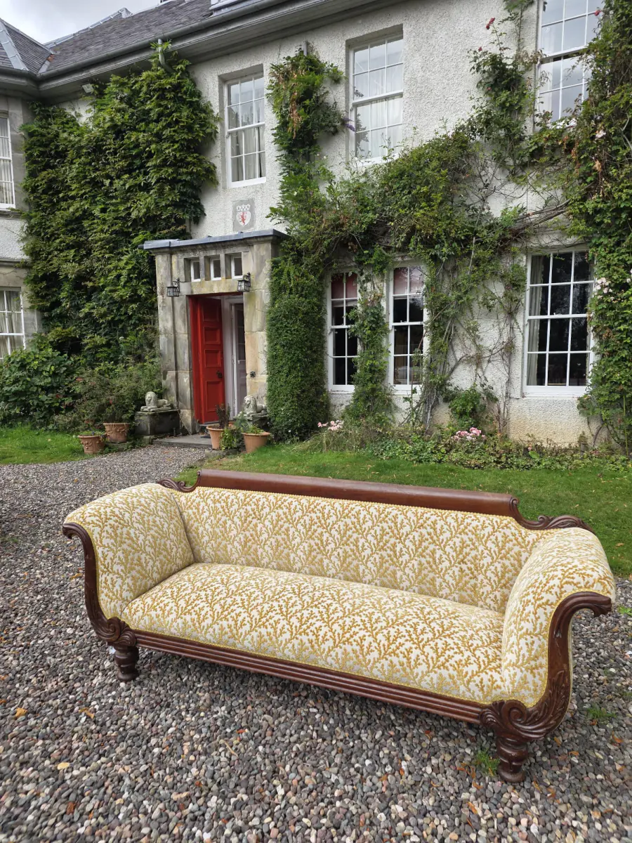 A Regency mahogany scroll-arm sofa reupholstered in a gold and cream fern print, on the gravel outside a country house